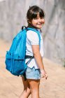 Visão traseira da criança em idade escolar com mochila no pavimento olhando para a câmera sobre o ombro sob a luz solar — Fotografia de Stock