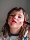 Enfant avec applicateur maquillage visage avec des produits cosmétiques assortis dans la maison regardant la caméra — Photo de stock