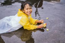 Affascinante bambino etnico in impermeabile che gioca con anatre di plastica che si riflettono nella pozzanghera increspata nel tempo piovoso — Foto stock