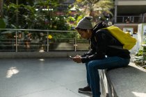 Contenuto hipster afroamericano maschile con zaino seduto sul bordo di pietra in strada e messaggistica sul cellulare e ascoltare musica su auricolari — Foto stock