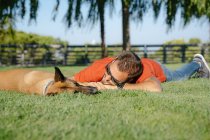 Niveau du sol du jeune homme barbu dans des lunettes de soleil couché sur la prairie contre chien de race dans le parc le jour de l'été — Photo de stock