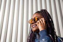 Cheerful stylish young African American female with Afro braids wearing trendy jacket and sunglasses speaking on mobile phone while standing near wall of urban building — Stock Photo