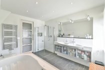 Современный санузел с ванной против душевой кабины и косметикой на полке в светлом доме — стоковое фото