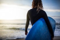 Vue arrière d'une jeune femme méconnaissable debout sur la rive avec une planche de surf avant d'entrer dans la mer au coucher du soleil sur la plage des Asturies, Espagne — Photo de stock