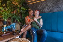 Contenu jeune femme tatouée avec mohawk et boisson embrassant petite amie lesbienne tout en se regardant sur le canapé dans la maison — Photo de stock