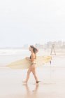 Вид сбоку на счастливую молодую спортсменку в купальниках с доской для серфинга, смотрящую на песчаное побережье против бурного океана — стоковое фото