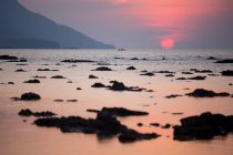 Wide coast with seaweeds of rippling sea against hill and red sun at sundown in Malaysia — Stock Photo