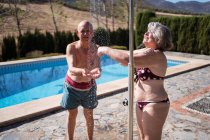 Hombre sin camisa de pie cerca de la vieja esposa positiva y tomar una ducha en la piscina en el soleado día de verano en el patio trasero - foto de stock