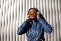 Happy young African American female with Afro braids dressed in blue jacket and stylish sunglasses enjoying music through earphones while chilling in sunlight against striped wall — Stock Photo