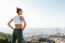 Back view of fit female athlete with hands on waist admiring summer town in sunlight — Stock Photo