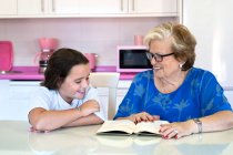 Nonna concentrata in abiti casual e occhiali da vista seduta a tavola e libro di lettura con allegra nipote in cucina a casa — Foto stock