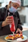Етнічний чоловічий кухар у стерильній масці прикрашає смачні віденські вафлі з збитими вершками з пляшки на кухні ресторану. — стокове фото