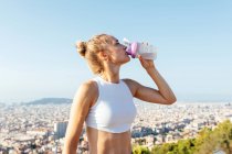 Slim female athlete with hand on hip and closed eyes drinking water from bottle during break from workout in town — Stock Photo