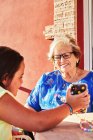 Nieta feliz sentada en la mesa y mostrando fotos en el teléfono inteligente a la abuela sonriente mientras está sentada en la terraza en un día soleado - foto de stock