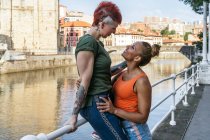 Вид сбоку на веселую молодую гомосексуальную женщину, обнимающую татуированную подружку с ирокезом, глядя друг на друга на канале в городе — стоковое фото