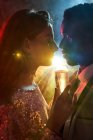 Vista lateral do casal de culturas com vidro em champanhe no momento do beijo contra o raio de luz brilhante olhando um para o outro durante a festa — Fotografia de Stock