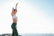 Ajuste jovem atleta feminina em sportswear exercitando com braço levantado enquanto olha para longe sob o céu claro na cidade ensolarada — Fotografia de Stock