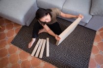 Mesa de montaje femenina étnica atenta joven en alfombra ornamental contra sofá en la sala de casa de luz - foto de stock