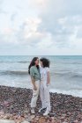 Young lesbian girlfriends in casual wear embracing while looking at each other on ocean coast under cloudy sky — Stock Photo