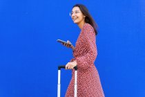 Seitenansicht einer positiven Frau im langen roten Kleid, die mit Gepäck unterwegs ist, während sie tagsüber auf der Straße mit dem Smartphone gegen eine blaue Wand surft — Stockfoto