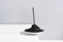Tenedor de acero inoxidable en un tazón lleno de espagueti negro con tinta de sepia sobre mesa blanca en estudio sobre fondo gris - foto de stock