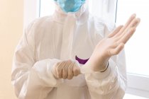 Médico masculino anónimo de cosecha en traje PPE que se pone guantes de látex mientras se prepara para el trabajo durante la pandemia de coronavirus en el hospital - foto de stock