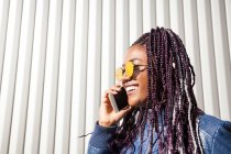 Joven mujer afroamericana alegre y elegante con trenzas afro con chaqueta de moda y gafas de sol hablando por teléfono móvil mientras está de pie cerca de la pared del edificio urbano - foto de stock