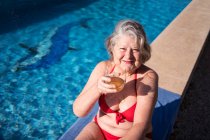 Hochaufragende Seniorin im Bikini lacht fröhlich, während sie am Pool mit Getränken chillt — Stockfoto
