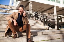 Стильный молодой человек в очках и повседневной одежде, сидящий на лестнице — стоковое фото