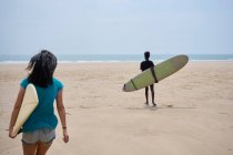 Vista posterior de jóvenes surfistas femeninas multiétnicas con tablas de surf hablando mientras pasean en la orilla arenosa y mirando y entre sí - foto de stock