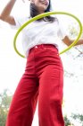 Из-под урожая неузнаваемая весёлая девушка-подросток в красных джинсах крутит хула-хуп во время свободного времени в парке — стоковое фото