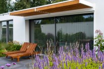 Деревянные шезлонги, размещенные под крышей современной жилой виллы со стеклянными стенами на заднем дворе, украшенные цветущими цветами в солнечный день — стоковое фото