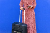 Gestutzte, bis zur Unkenntlichkeit verkleidete Frau im langen roten Kleid, die mit Gepäck auf der Straße steht, während sie tagsüber eine Nachricht auf dem Smartphone an eine blaue Wand schreibt — Stockfoto