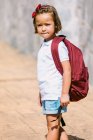 Seitenansicht eines Schulkindes mit Rucksack auf dem Bürgersteig, das im Sonnenlicht in die Kamera blickt — Stockfoto