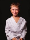 Lindo chico en kimono de karate riendo felizmente con la boca abierta en el estudio sobre fondo negro - foto de stock