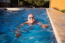 Relaxed senior male in glasses swimming in transparent clean pool water while relaxing in hot summer day — Stock Photo