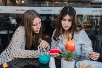 Migliori amiche con bicchieri di bevande rinfrescanti che navigano sul cellulare a tavola nel caffè urbano — Foto stock