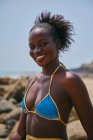 Бічний погляд на вміст молодої афроамериканської жінки в купальнику, яка сидить на валуні, дивлячись на камеру. — стокове фото