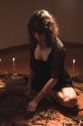 Enigmatique sorcière en robe noire assise faisant cercle avec des branches et des bougies allumées pendant le rituel spirituel dans la chambre noire — Photo de stock