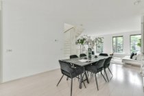 Interior of dining zone with large table with bouquet of flowers and chairs in modern apartment in daytime and in the background the living room and bright windows. — Stock Photo