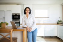 Заколдованная счастливая зрелая женщина-фрилансер, просматривающая в Интернете нетбук, работающий над новым проектом, стоя, глядя в камеру на столе на кухне дома — стоковое фото