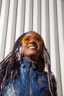 De baixo feliz jovem afro-americano feminino com tranças afro vestidas com jaqueta azul e óculos de sol elegantes desfrutando de música através de fones de ouvido enquanto arrefece na luz solar contra a parede listrada — Fotografia de Stock