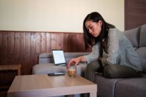 Вид збоку концентрованої молодої етнічної жінки автора з кавою, що сидить на дивані на мобільному телефоні та нетбуці вдома — стокове фото