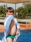 Adorable petit enfant avec des cheveux mouillés essuyant le corps avec une serviette tout en se tenant près de la piscine par une journée ensoleillée en été et en regardant la caméra — Photo de stock