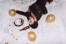 Пьяный смеющийся мужчина в разбитом торте, лежащем рядом с пустыми бутылками пива и воздушными шарами с закрытыми глазами — стоковое фото