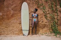 Vorderansicht einer afroamerikanischen Sportlerin, die mit einem Surfbrett von einem Strandabschnitt wegschaut und vor einem Lehmfelsen mit Pflanzen an der Seite — Stockfoto