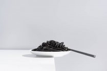 Tenedor de acero inoxidable en un tazón lleno de espagueti negro con tinta de sepia sobre mesa blanca en estudio sobre fondo gris - foto de stock
