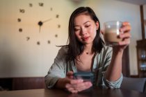 Молода етнічна жінка зі склянкою кави, що серфінгує Інтернет на мобільному телефоні за столом у домашній кімнаті — стокове фото