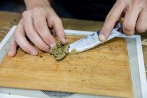 Crop unrecognizable male with knife mashing dried cannabis plant piece on wooden board in workspace — Stock Photo