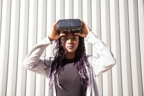 Возбужденная молодая афроамериканка в наушниках VR развлекается и играет в виртуальную игру против серой полосатой стены — стоковое фото